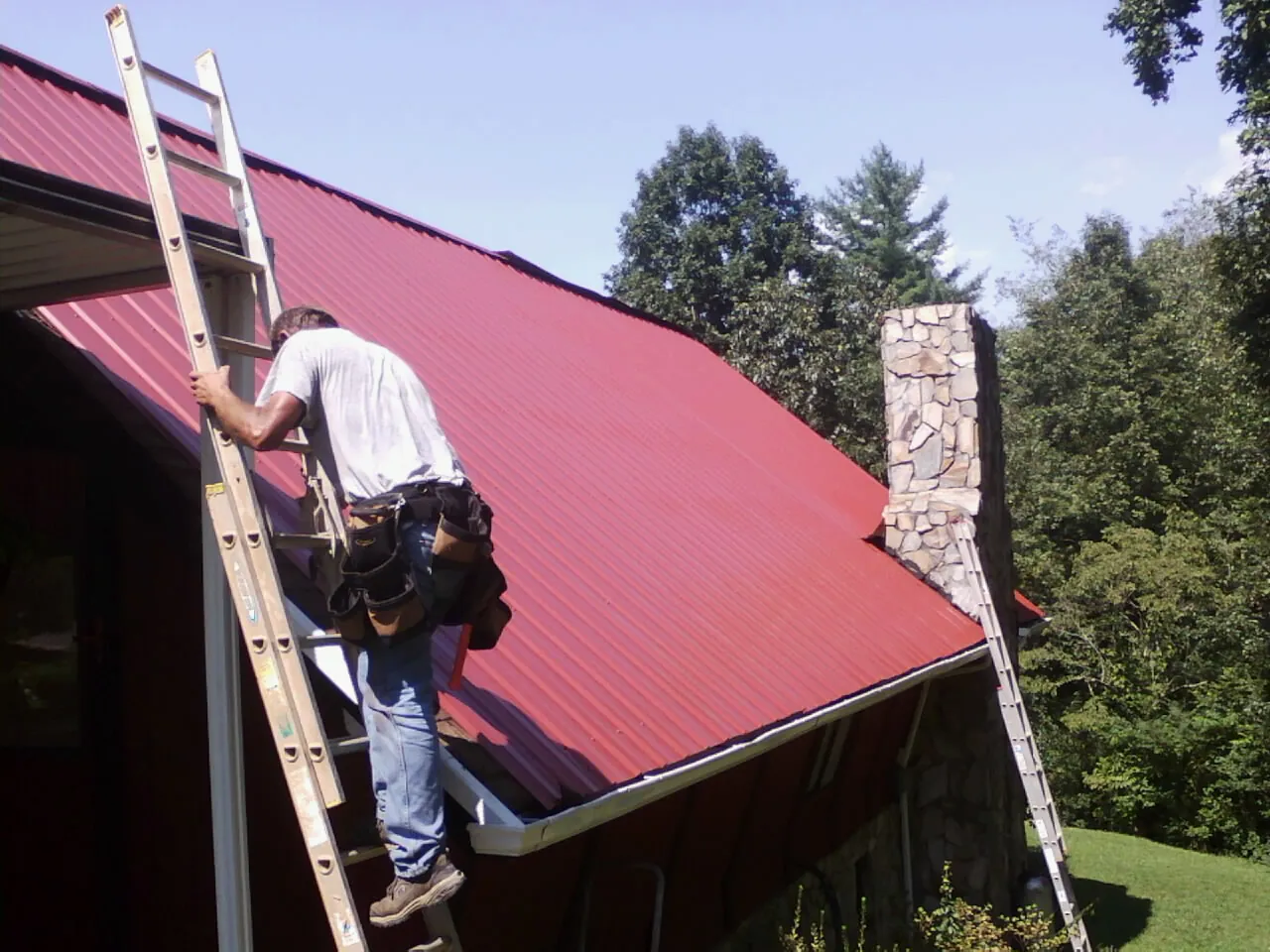 Roofer on a ladder beside a red metal roof and stone chimney — Soffit & Fascia Repair by Roofing Brothers in Colquitt